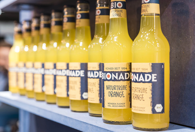 Aufgereihte Bionade Flaschen der Sorte „Naturtrübe Orange“ auf einem blauen Regalbrett